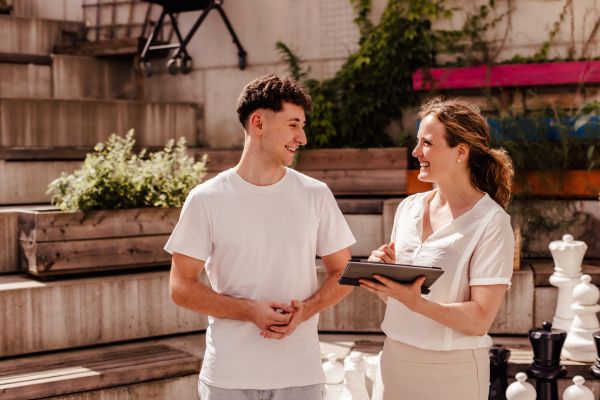 Beratungssituation: eine junge Frau sitzt lächelnd gegenüber einem Betreuer in einem Büro. Auf dem Schreibtisch liegen Unterlagen und im Hintergrund ist ein Bild mit Sonnenblumen zu sehen.