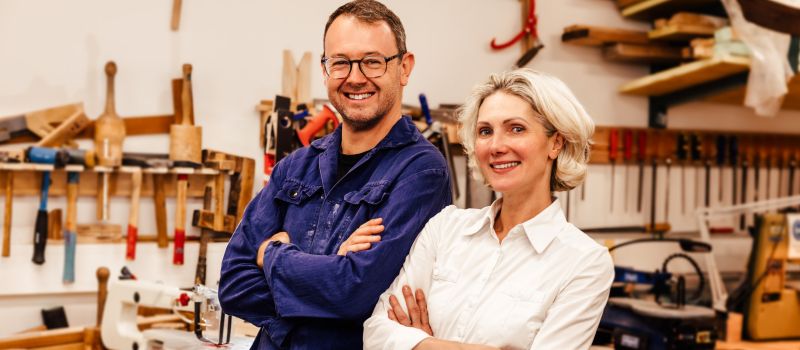 Ein Mann in Arbeitskleidung und eine Frau in Bluse stehen Rücken an Rücken in einer Holzwerkstatt und lachen.