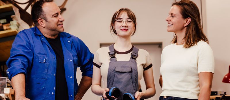 Drei Personen stehen in einer Holzwerkstatt. In der Mitte steht eine Jugendliche mit Arbeitskleiung und blickt selbstbewusst in die Kamera. Rechts und links stehen ihr zugewandt lächelnd ein Mann in Arbeitskleidung und eine Frau in Jeans und TShirt.